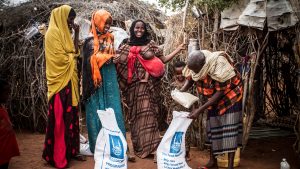 Tre kvinnor skrattar, en bebis hänger på ena kvinnans mage i en sjal, ett barn ler och en man packar upp saker ur en stor säck