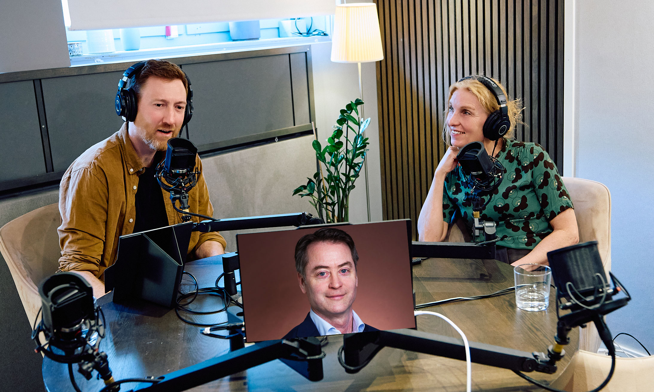 En man och en kvinna i en poddstudio med en man på skärm