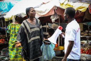 Mörkhyad kvinnai svart klänning argumenterar med mörkhyad man i vit T-shirt och megafon på afrikansk marknad